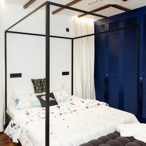 Photo of a bedroom framed by a slim black steel four-poster around a white bed, with a full wall of navy louvered wardrobe shutters and black bar handles