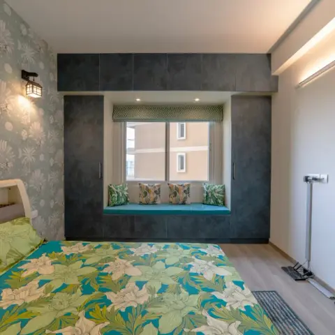 Photo of a bedroom with large-scale palm-print wallpaper behind the bed, a concrete-textured wardrobe wrapping a window seat cushioned in teal