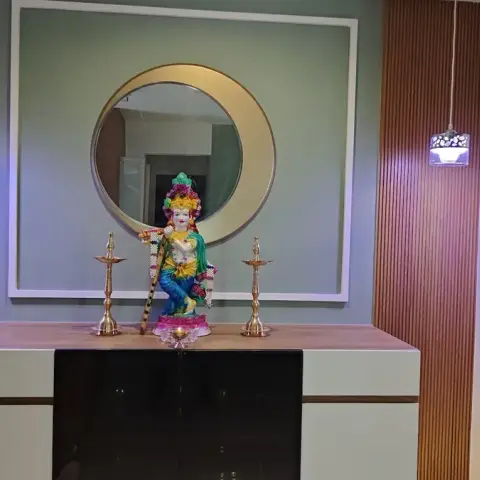 Photo of a home pooja unit: a garlanded Krishna idol between two tall brass diyas on a wood-topped white console