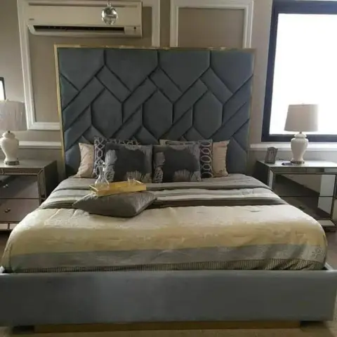 Photo of a master bedroom with a tall channel-tufted grey velvet headboard bordered in gold trim, mirrored bedside tables