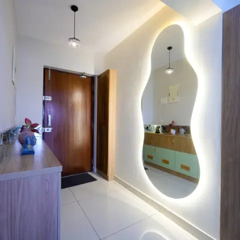 Photo of an entry foyer where a backlit free-form mirror is cut into a walnut partition facing the main door, reflecting a mint-green sideboard