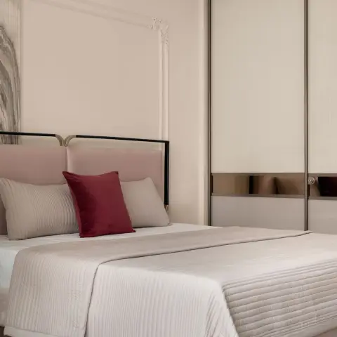 Photograph of a bedroom corner: a blush velvet headboard held in a black-and-brass frame, cream quilted bedding with one burgundy velvet cushion