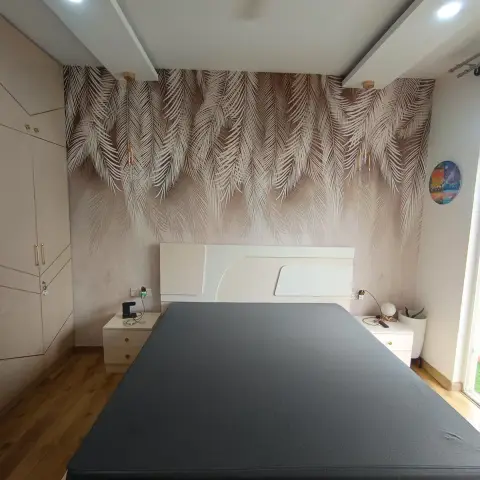 Photograph of a bedroom with a blush palm-frond mural covering the headboard wall, slim brass pendant drops hanging either side of a white curved headboard
