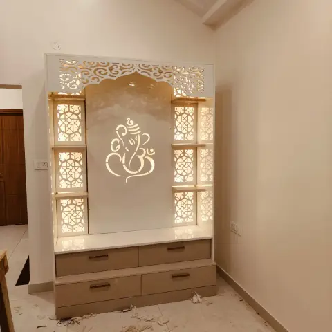Photograph of a freestanding white-and-beige pooja unit - backlit CNC jaali side panels and a cut-out Ganesha motif over a marble platform