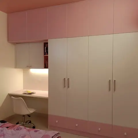 Pink and white kids' bedroom with a built-in study desk nook under wall cabinets beside a full-height wardrobe