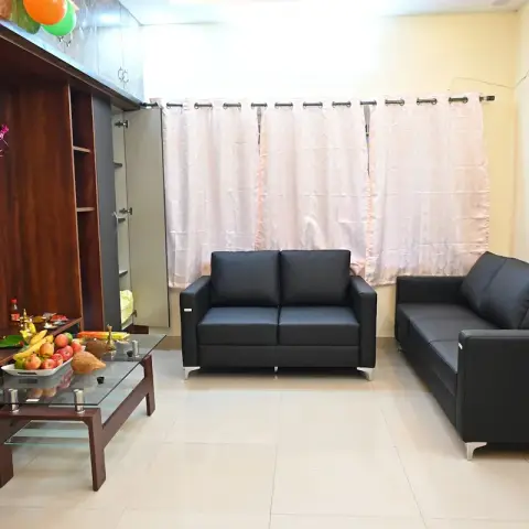 Plainly furnished living room set up for a housewarming puja, with garlanded deity photos and fruit offerings laid out on the dark wood wall unit and glass