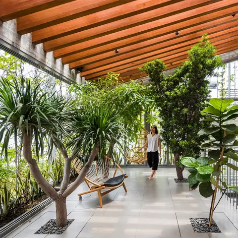 Plant-filled balcony corridor with a wood-slat ceiling, potted trees lining a stone floor, and a woven hanging swing chair.