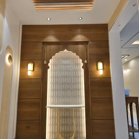 Pooja alcove with an arched, backlit textured tile niche framed by walnut wood panelling, a brass console table, and hanging temple bells.