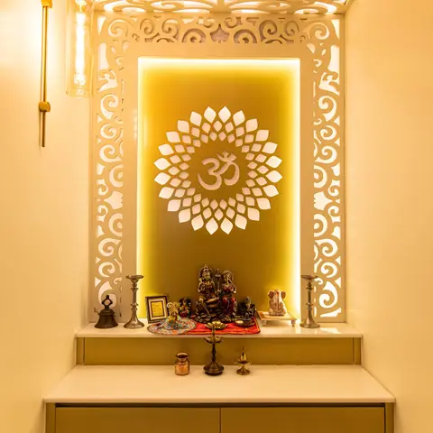 Pooja alcove with an ornate white CNC-cut jaali frame wrapping the wall and ceiling around a backlit Om medallion, brass idols on a marble ledge