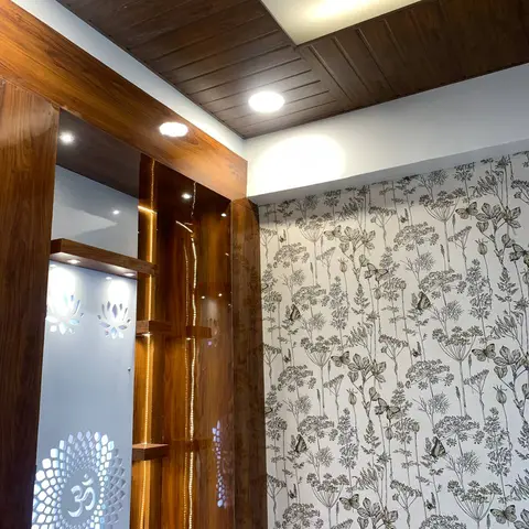 Pooja corner with a backlit lotus-and-Om cutout panel in a wood-laminate unit, set beside a wood slat ceiling and botanical butterfly wallpaper.