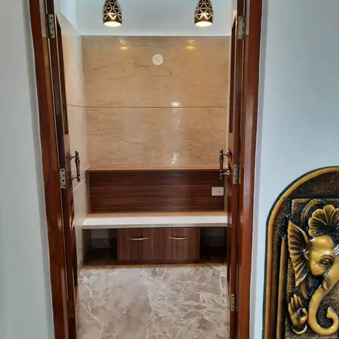 Pooja room seen through open wood-framed doors, with marble-clad walls, a built-in altar shelf with drawers, two pendant lights