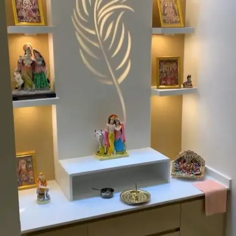 Pooja room with a backlit CNC-cut peacock-feather wall panel, symmetrical display shelves of framed deities, and a raised platform with Radha-Krishna idols