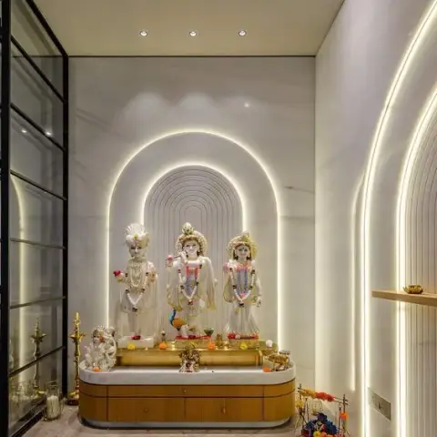 Pooja room with a backlit marble arch niche housing deity idols, a fluted wood cabinet base, brass diyas, and a glass sliding door framing the space.