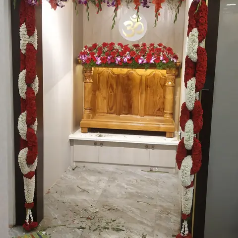 Pooja room with a carved wooden mandir unit and a backlit Om symbol, framed by fresh floral garlands and petals from a housewarming ceremony.