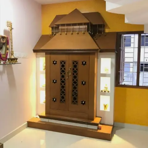 Pooja room with a temple-shaped wooden mandir unit featuring star-cutout jaali doors and lit display niches, set against a mustard-yellow textured wall.