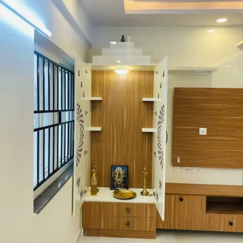 Pooja unit built into the living wall next to the TV panel — oak-laminate back with white CNC-cut jaali side shutters folded open, a stepped white gopuram top