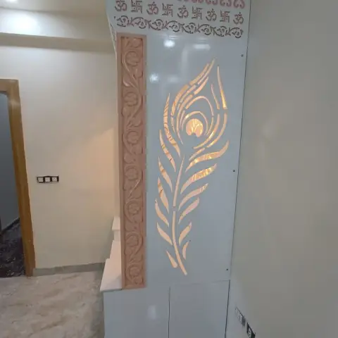 Pooja unit corner with a backlit laser-cut peacock feather panel beside a hand-carved floral wood pillar, Om and swastik motifs along the top border.