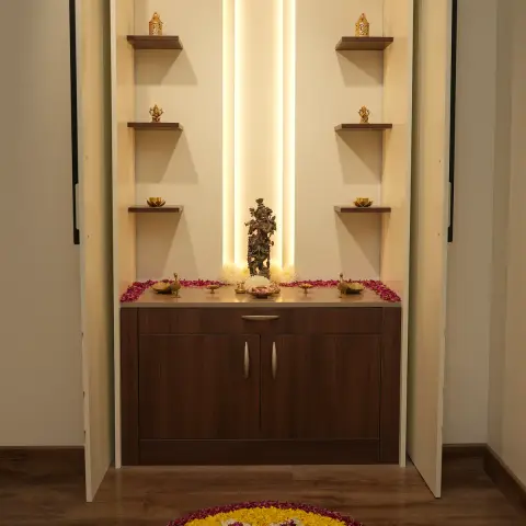 Pooja unit set in a niche behind full-height ivory shutters, a curved warm LED profile framing a brass Krishna idol on a wenge counter edged with rose petals