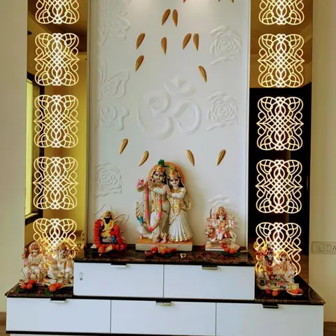 Pooja unit with backlit gold jaali side panels, a carved Om and floral relief wall, and marble Radha-Krishna and Ganesh idols on a white cabinet.