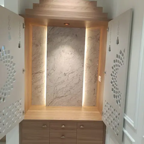 Pooja unit with open CNC-cut jaali doors revealing a backlit marble niche and tiered wood pediment above a walnut base cabinet with brass hardware.