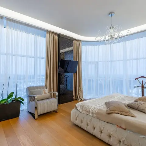 Primary bedroom with a curved floor-to-ceiling glass wall, sheer and beige drapes, and a tufted upholstered bed