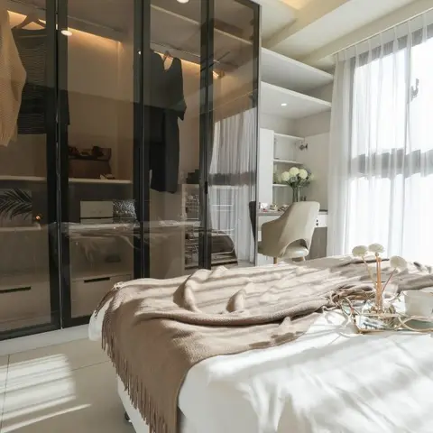 Primary bedroom with a glass-fronted walk-in wardrobe and a sunlit dressing nook beside sheer curtains.
