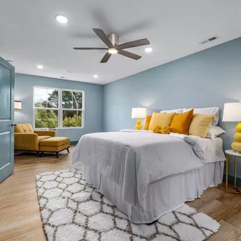 Primary bedroom with soft blue walls, a mustard velvet armchair and ottoman by the window, a wood ceiling fan