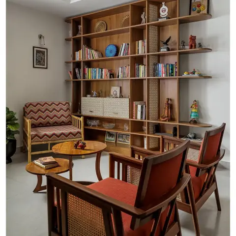Reading corner where a floor-to-ceiling teak shelf of books, brass plates and terracotta figures faces two cane-backed teak armchairs in rust wool and an
