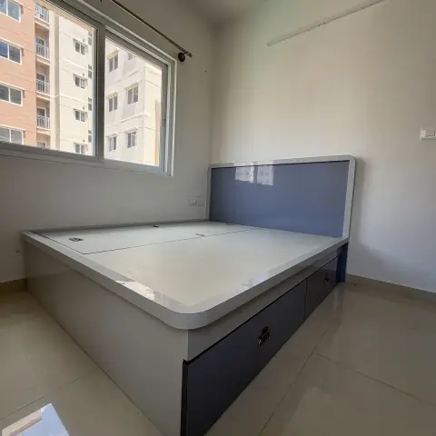 Real bedroom handover shot with a glossy grey-and-navy storage bed frame with drawer units, bare walls, and a single tube light fixture