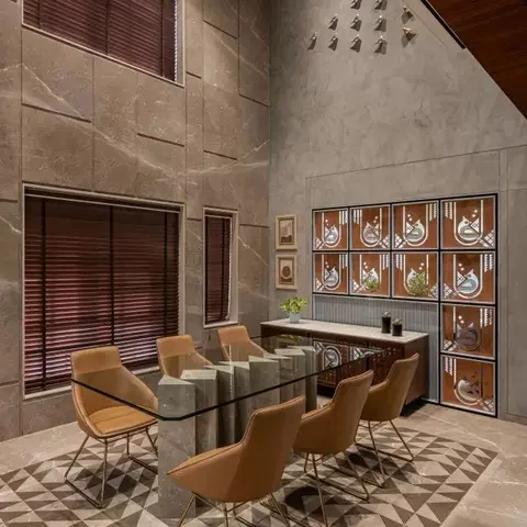 Real double-height dining room with marble-clad walls, a glass-top table on a sculptural stone base, tan leather chairs with gold legs, a brass chandelier