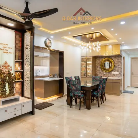 Real open-plan apartment showing an ornate white-and-wood pooja unit beside an open kitchen, a floral-upholstered dining set under a crystal chandelier