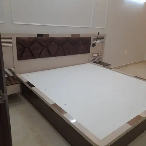 Real photo of a bedroom bed platform mid-handover with no mattress yet, a geometric tufted brown headboard, mirror-trimmed floating nightstands