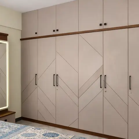 Real photo of a bedroom with a full-wall taupe wardrobe featuring a chevron groove pattern and a backlit LED mirror on a wood dresser beside the bed.