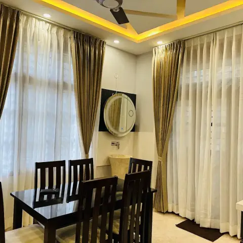 Real photo of a dining room with a dark wood table and chairs, floor-to-ceiling gold and sheer curtains, a cove-lit POP ceiling