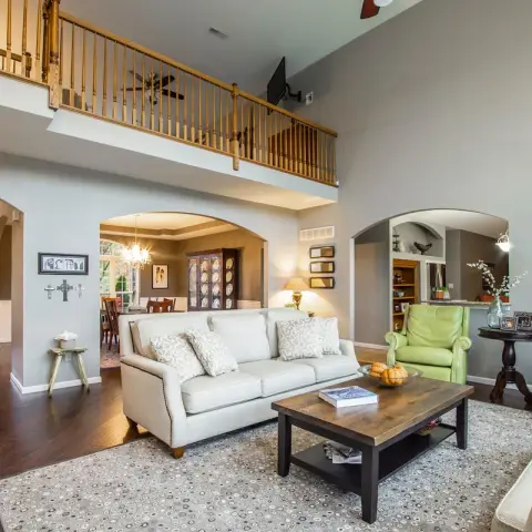 Real photo of a double-height living room with a wood-balustraded loft above, arched openings to a dining room and kitchen, cream sofas