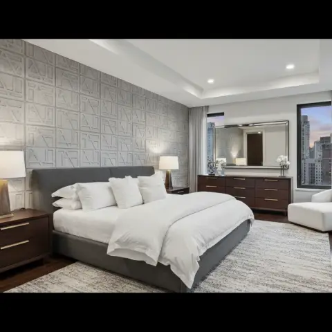 Real photo of a high-rise master bedroom with a geometric 3D-textured tile accent wall, a grey upholstered bed, a wood dresser with a large mirror