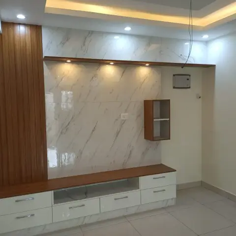 Real photo of a living room TV wall with marble-tile cladding, a fluted wood panel column, and a floating wood-and-white TV console under a cove-lit ceiling.