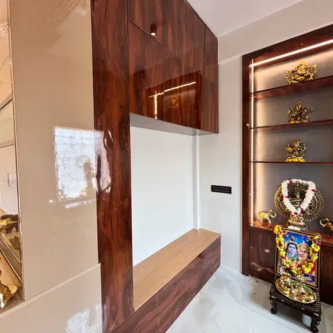Real photo of a pooja room with a backlit wood-clad brass idol display shelf and a glossy rosewood-veneer wrap-around wall