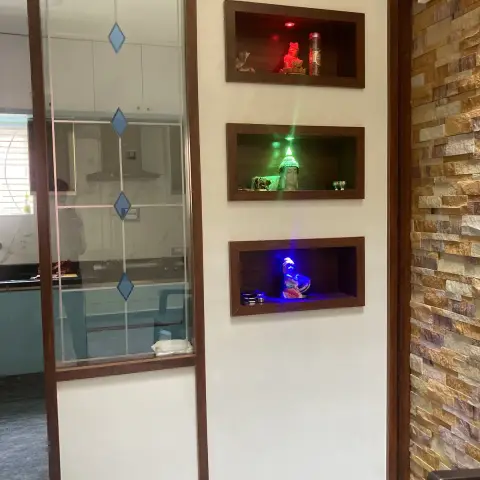 Real photo of a pooja wall with three dark-wood-framed backlit display niches holding deity idols
