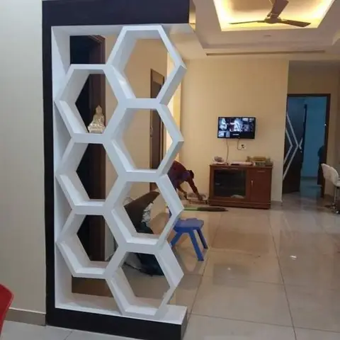 Real photo of a white hexagonal honeycomb partition unit dividing an entryway from a living room with a wall-mounted TV over a wood console and a cove-lit