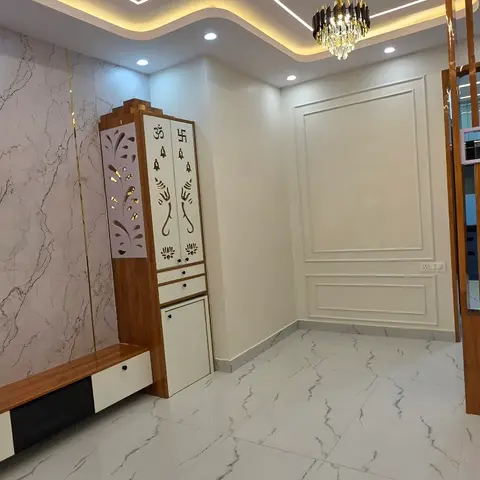 Real photo of an empty living room with a book-matched marble TV feature wall inlaid with brass strips, a compact wall-hung pooja unit with jaali-motif doors