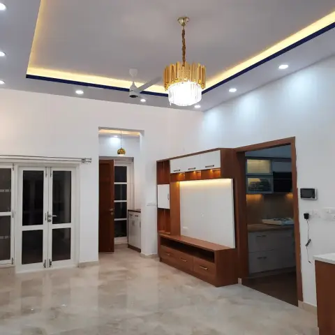 Real photo of an open-plan living space with a wood-and-white TV unit, a crystal-and-gold chandelier under a cove-lit ceiling, and glass French doors