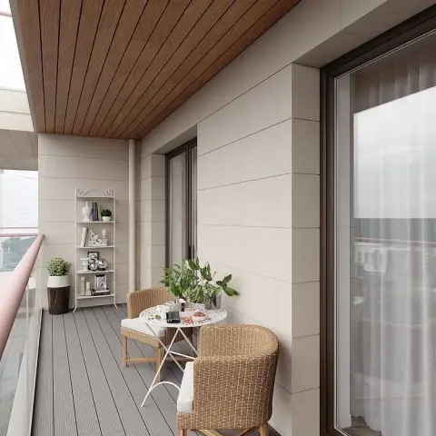 Render of a long enclosed apartment balcony with a teak-toned slatted soffit, grey composite decking and two wicker tub chairs pulled up to a white folding