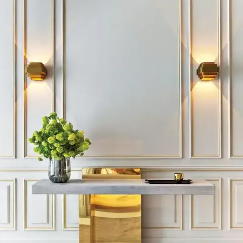 Render of an entrance console wall with white moulded wall panelling, gold wall sconces, and a brass-clad marble-top console.