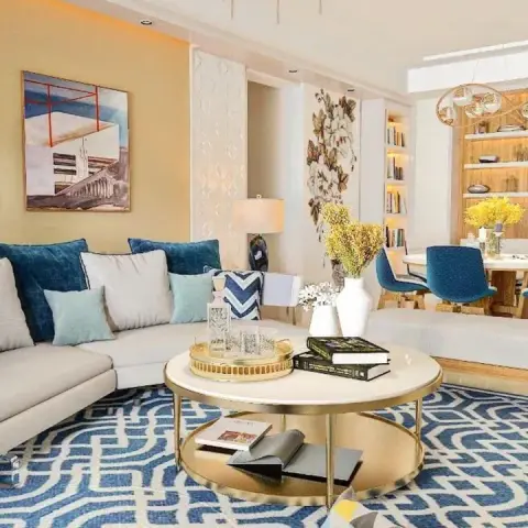 Render of an open living room with a white L-sofa piled with teal and indigo cushions over a blue maze-pattern rug, a two-tier brass coffee table