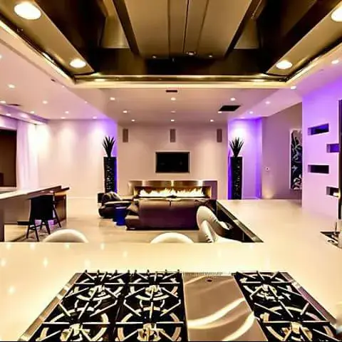 Render of an open-plan luxury space with a large kitchen island fitted with a professional gas range in the foreground and a fireplace lounge seating area