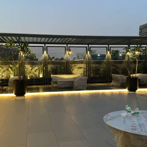 Rooftop terrace lounge under a slatted metal pergola with plastic-wrapped outdoor furniture, potted plants and pendant lights