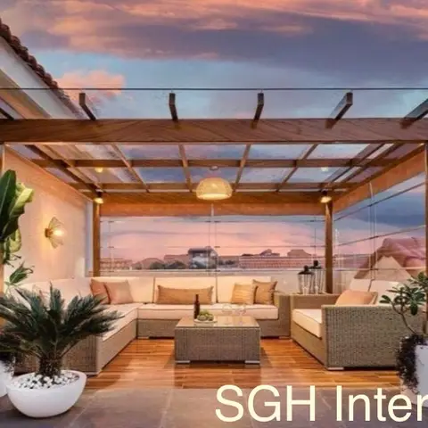 Rooftop terrace lounge under a wood-and-glass pergola with wicker sofa seating and a cluster of potted plants at sunset.