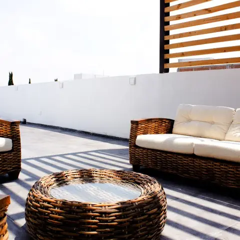 Rooftop terrace seating area with woven rattan sofa and armchair in cream cushions around a round rattan coffee table, screened by a timber slat pergola.