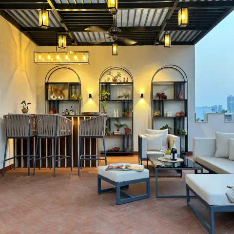 Rooftop terrace under a black steel pergola hung with brass cage lanterns, with woven bar stools at a lit counter and three arched black-frame shelving bays of